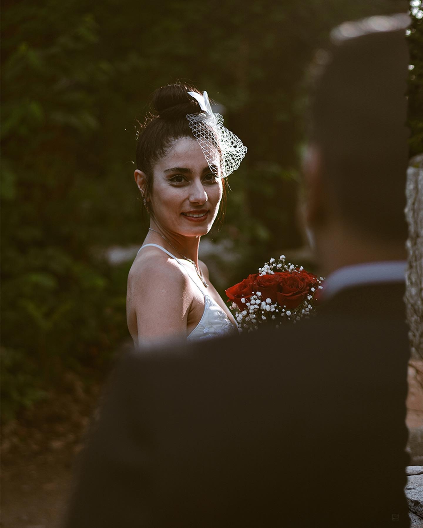 Bride with a birdcage veil and red roses smiling, framed past the blurred shoulder of the groom in the foreground