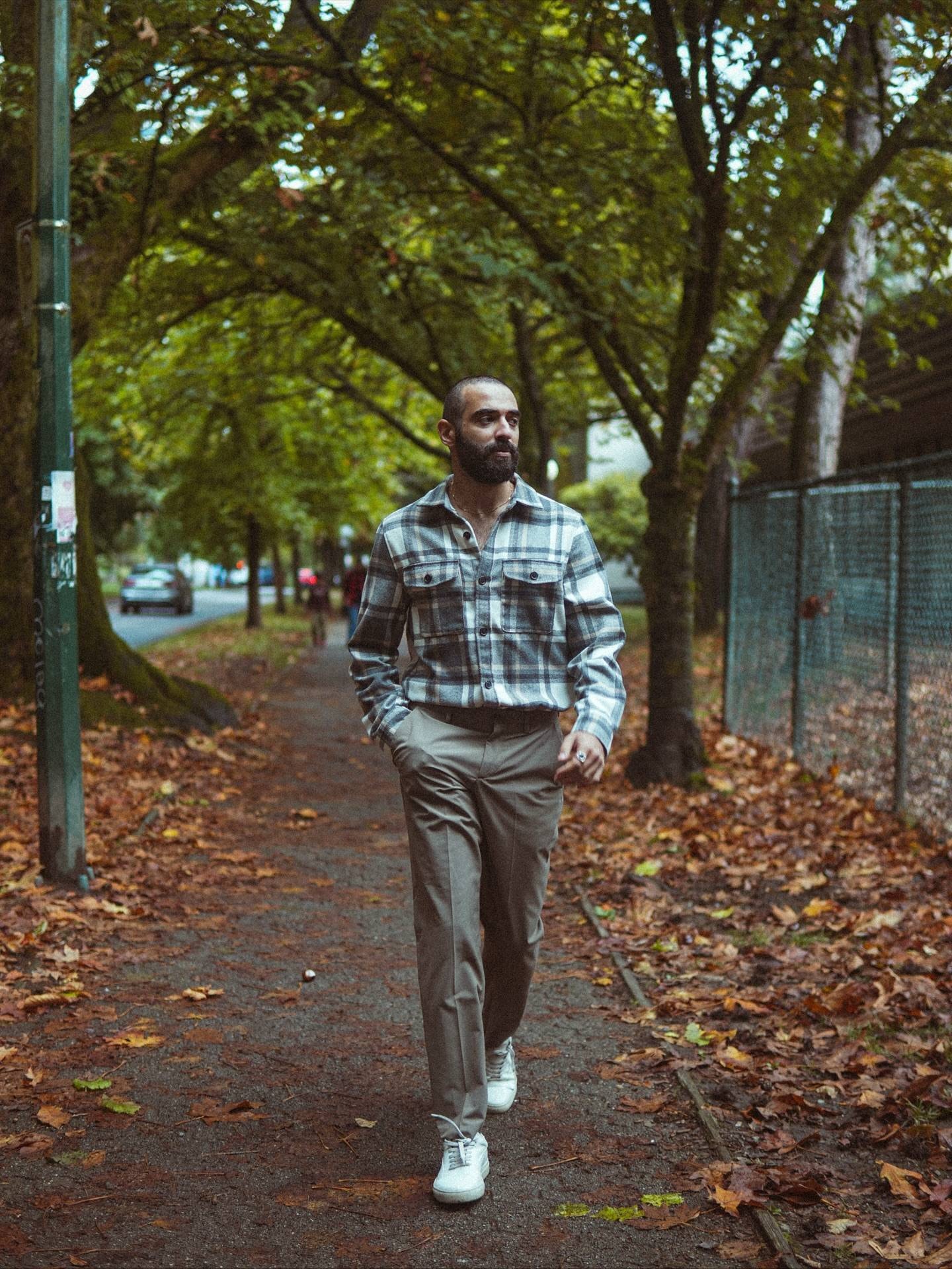 Bearded man in a grey plaid overshirt walking along a tree-lined path covered in autumn leaves