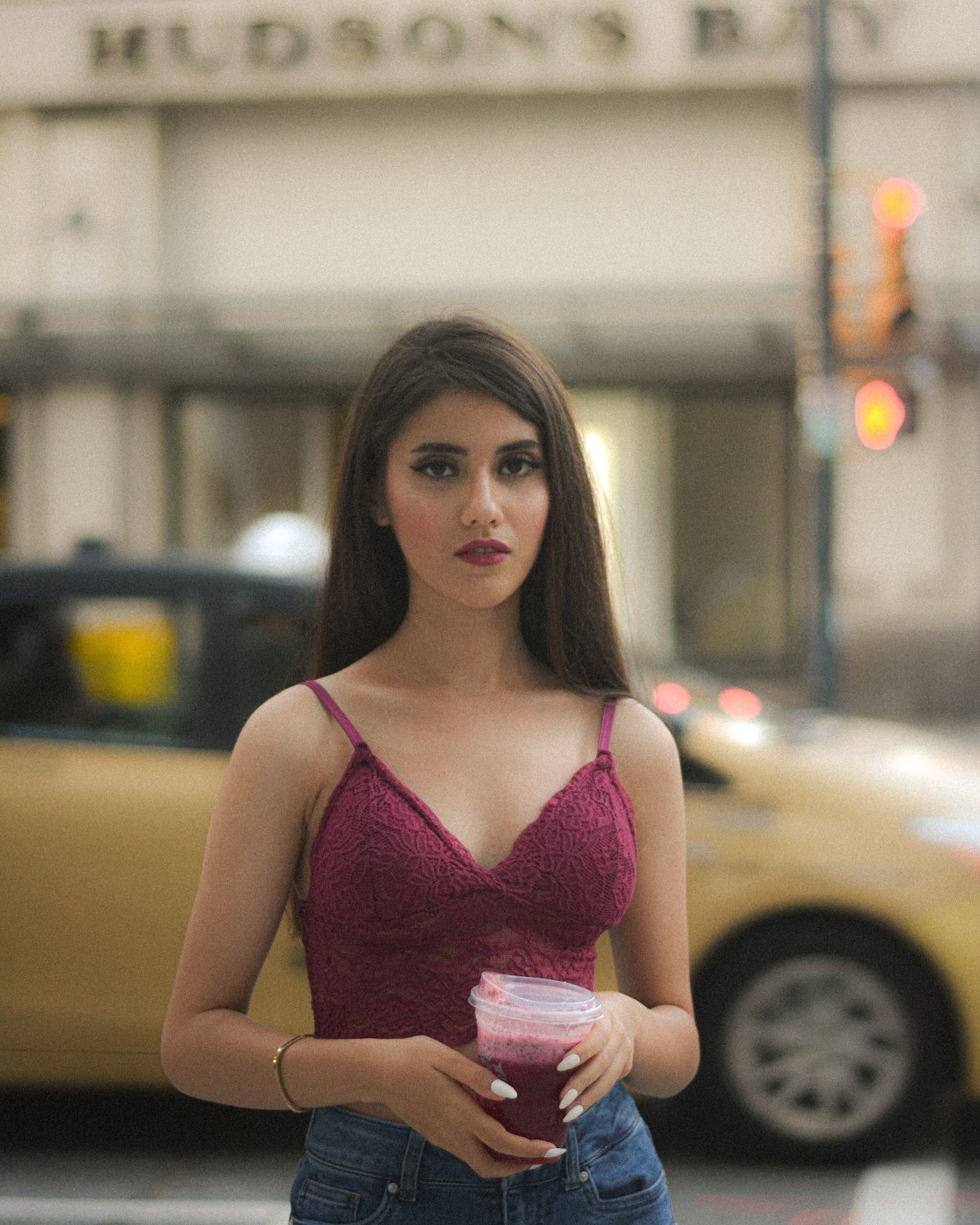 Woman in a burgundy lace top holding a berry drink on a downtown street, a yellow taxi passing behind her