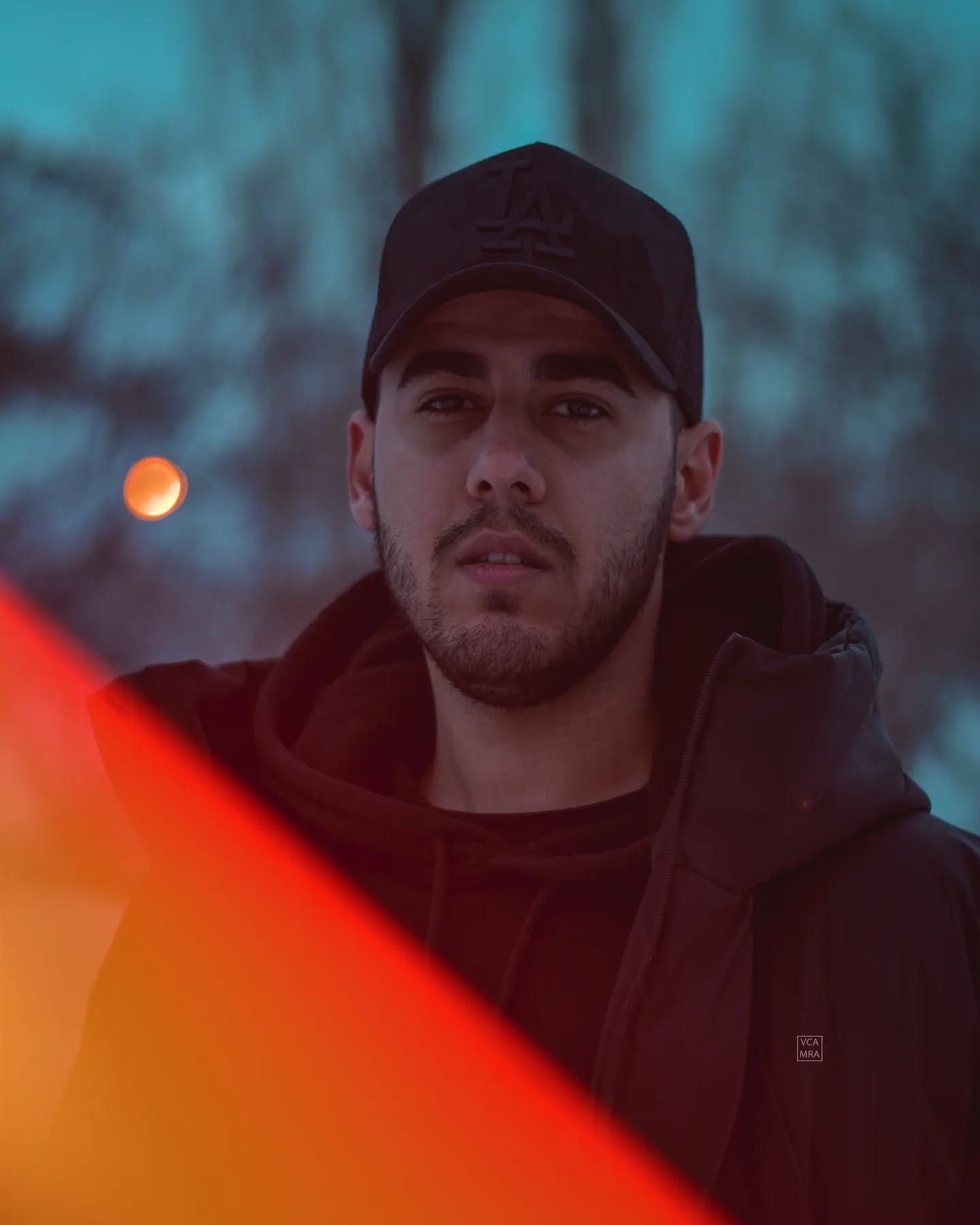 Man in a black cap and hoodie at blue hour, an orange light flare cutting across the lower corner of the frame