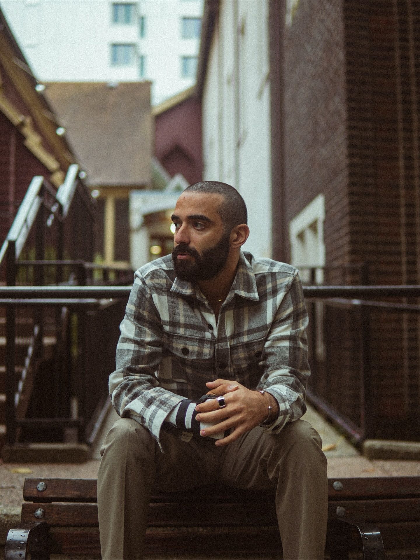 Bearded man in a grey plaid overshirt sitting on wooden steps holding a coffee cup, brick buildings behind him