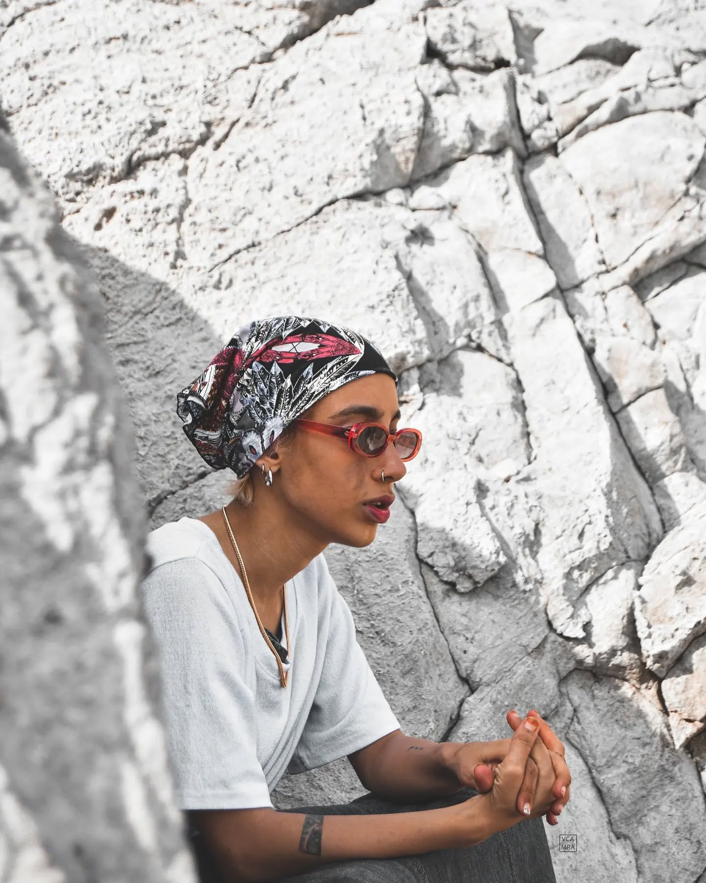 Woman in red oval sunglasses and a patterned head scarf sitting against a pale cracked rock face
