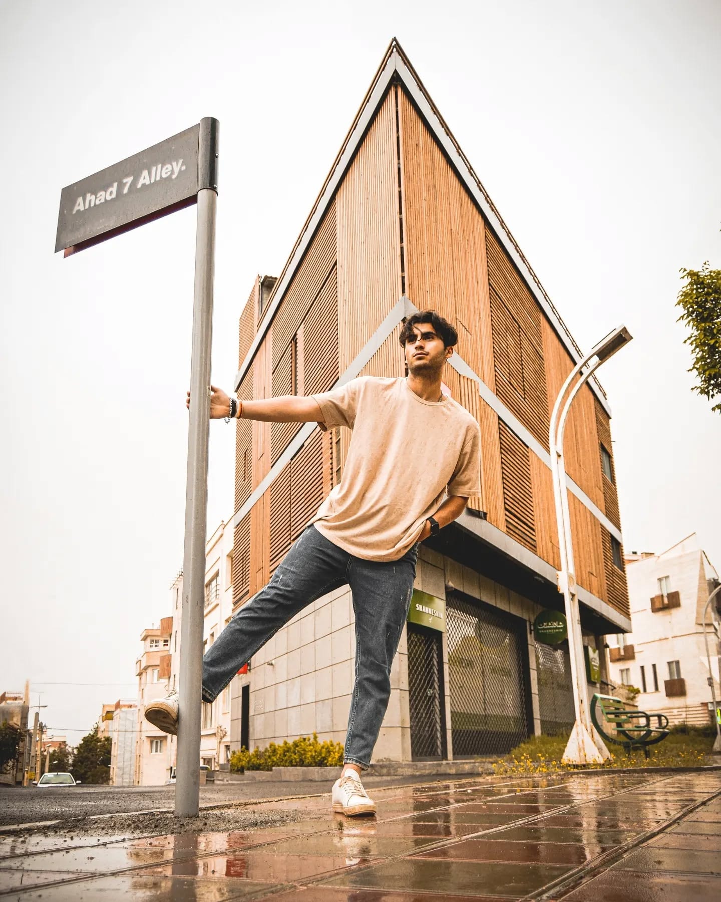 Young man in a beige t-shirt holding a street sign pole with one leg raised, a triangular wood-clad building behind him