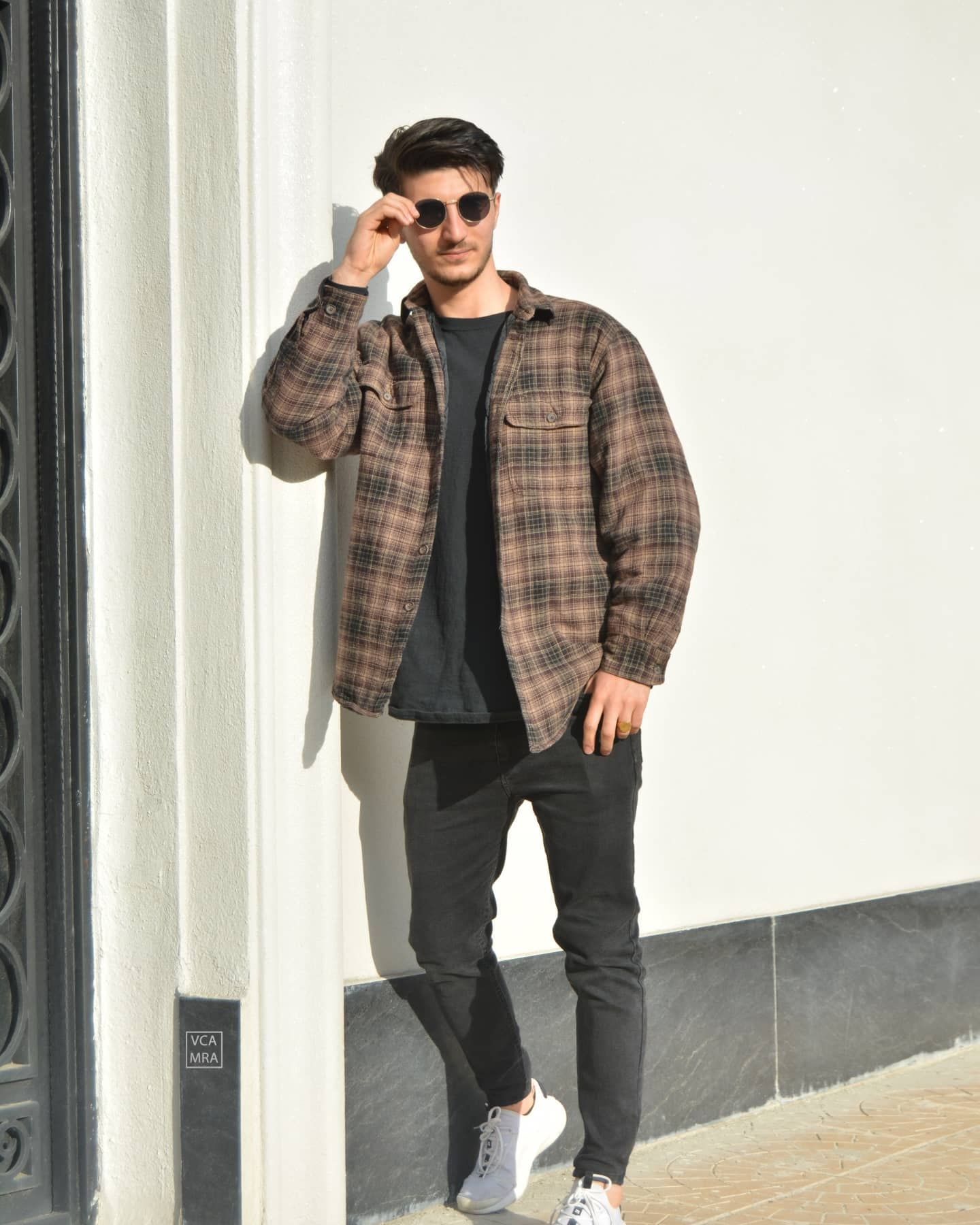 Man in a brown plaid shirt and black jeans leaning on a white wall, lowering his round sunglasses