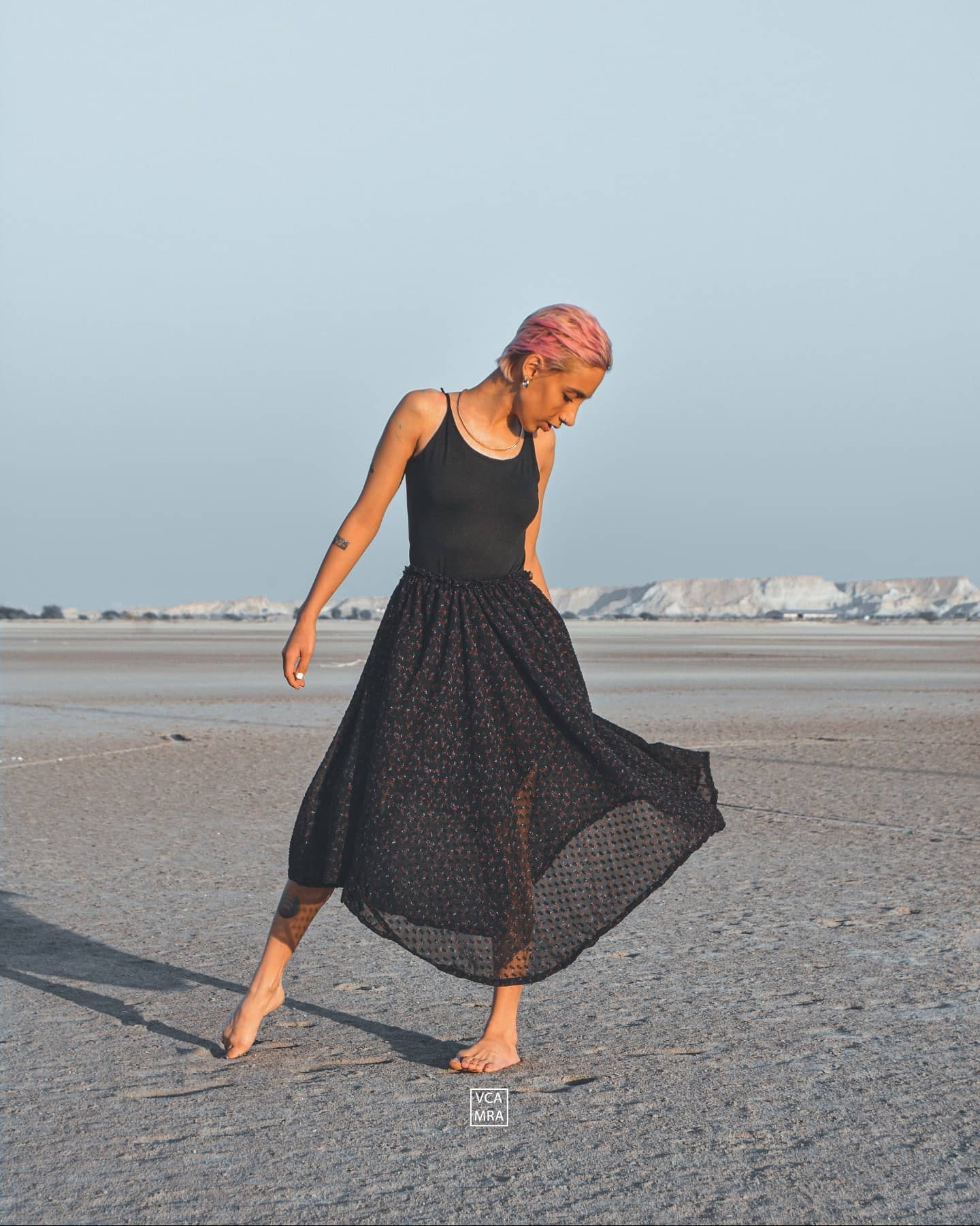 Woman with short pink hair turning barefoot on a salt flat, her black skirt swinging out
