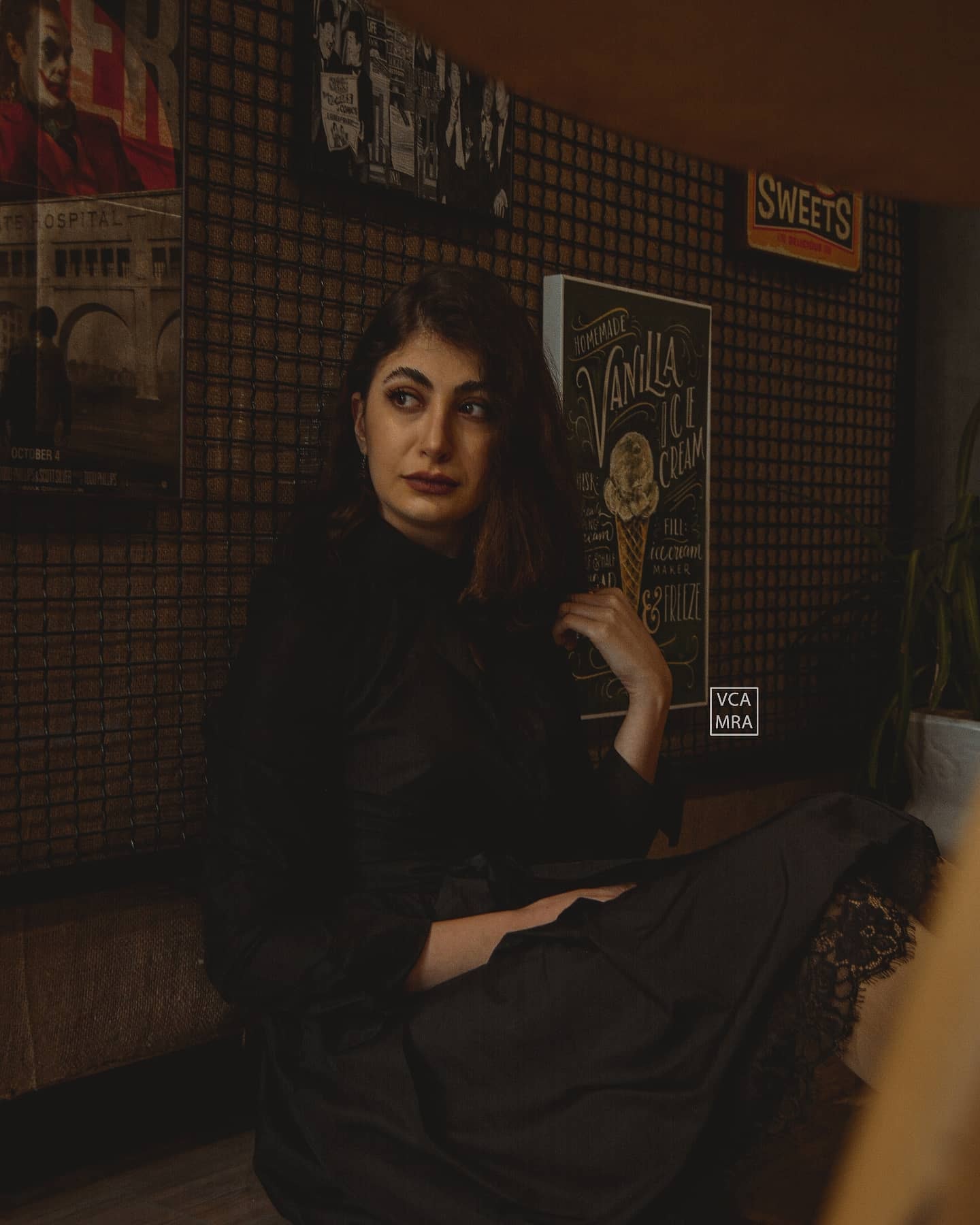 Woman in a black dress sitting in a dim café corner with posters and a vanilla ice cream sign on the wall
