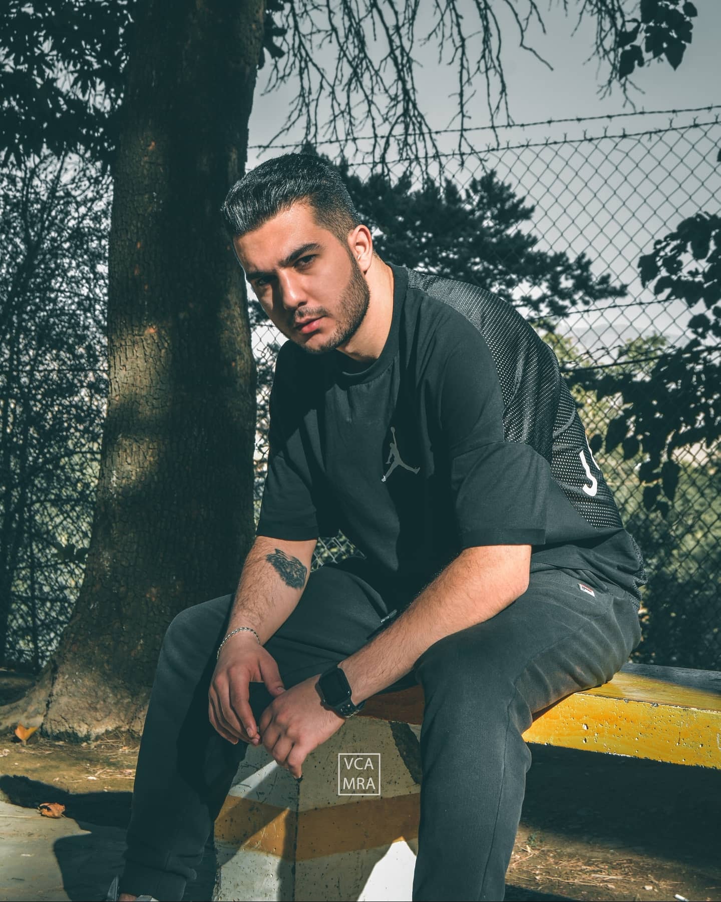 Man in a black t-shirt sitting forward on a yellow concrete ledge in front of a tree and chain-link fence