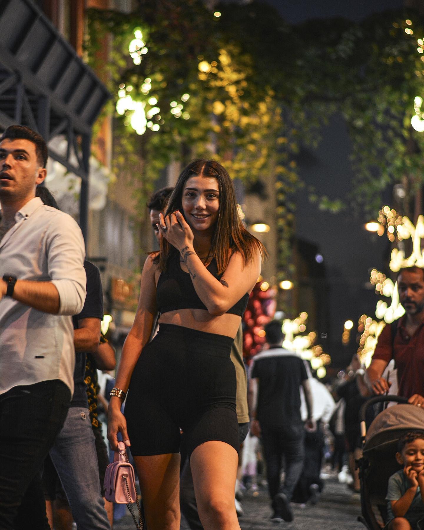 Smiling woman in black walking through a busy evening street lit with warm string lights