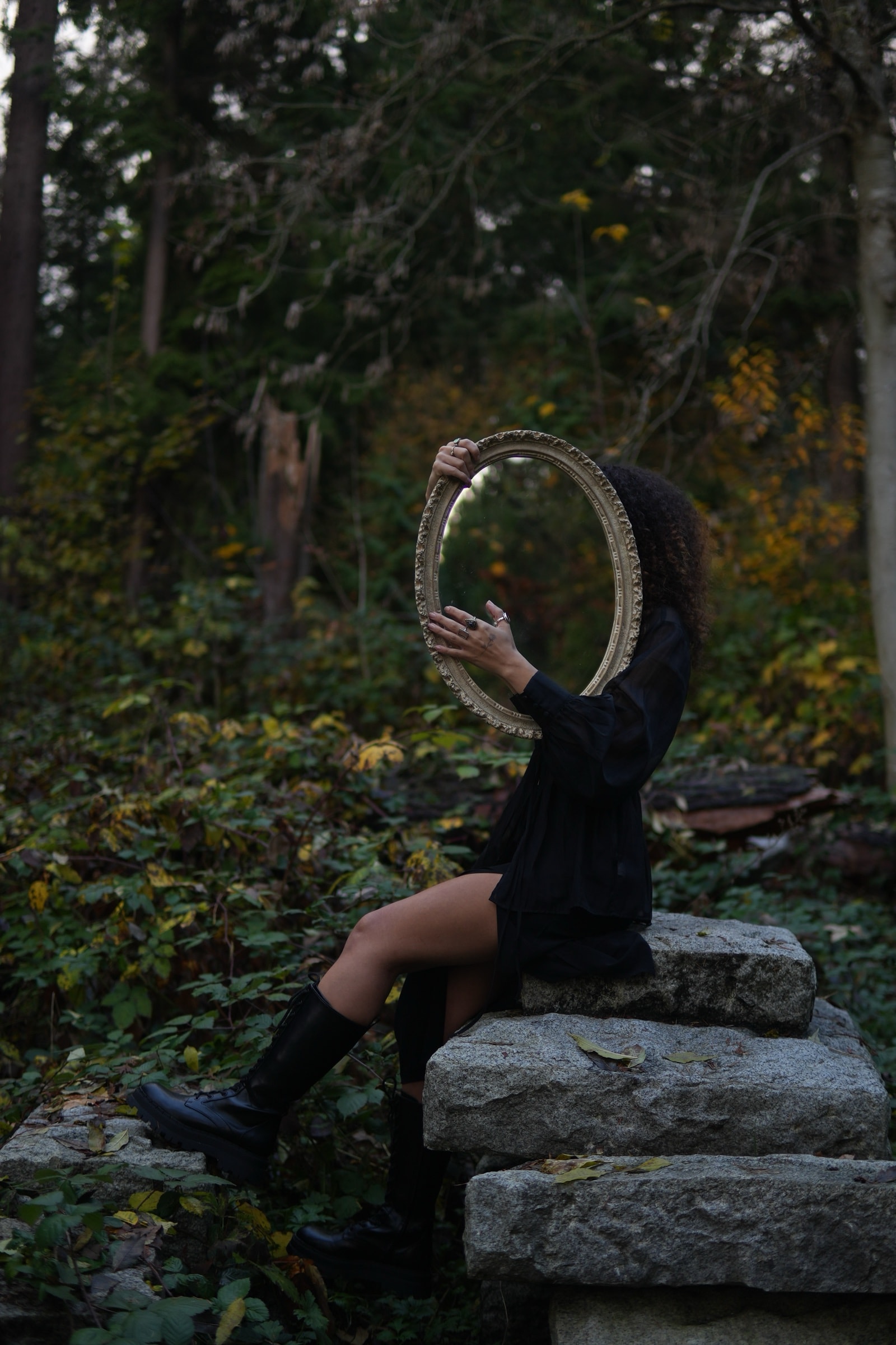 Woman in black sitting on mossy stone steps in a forest, holding an ornate oval mirror in front of her face