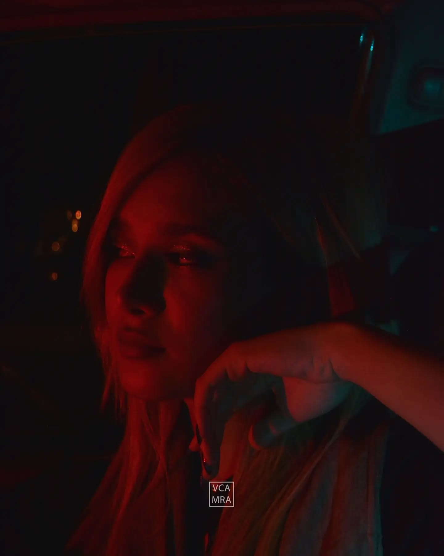 Blonde woman in deep red light inside a car at night, resting her head on her hand, city lights blurred behind