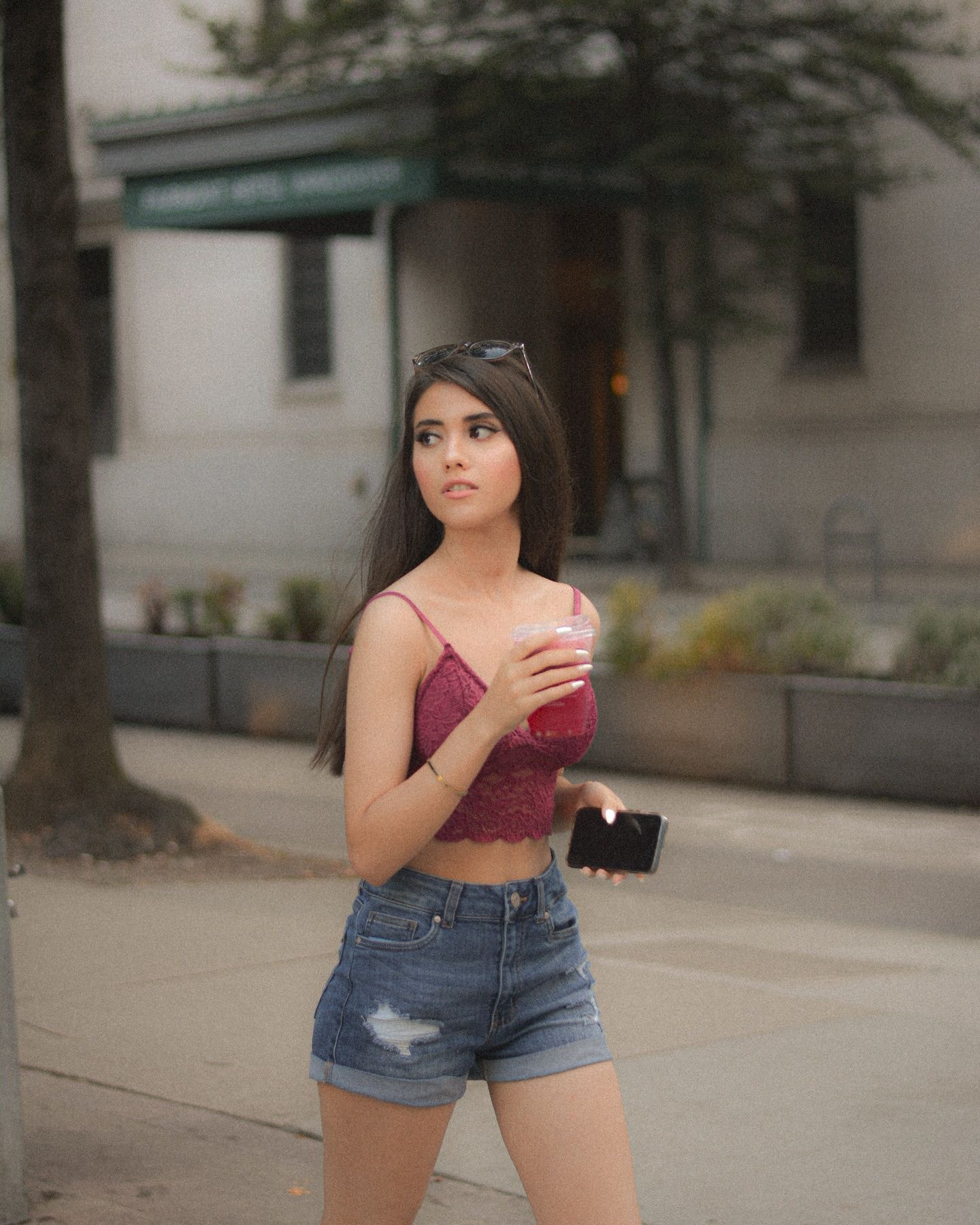 Woman in a pink lace top and denim shorts walking with a drink and phone, glancing over her shoulder