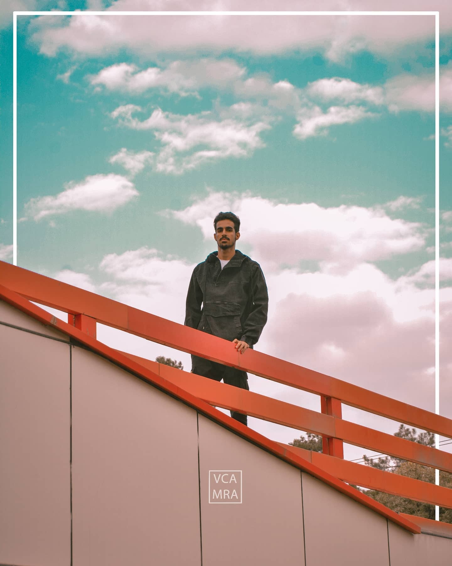 Man in a dark jacket standing behind an orange railing on an overpass under a cloudy teal sky