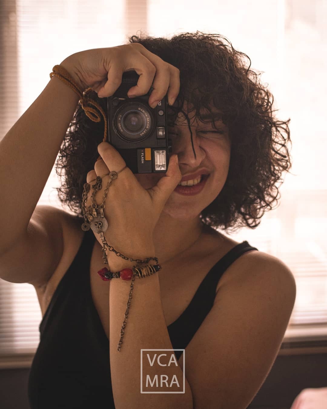 Laughing woman with curly hair holding a vintage film camera up to one eye in a sunlit room