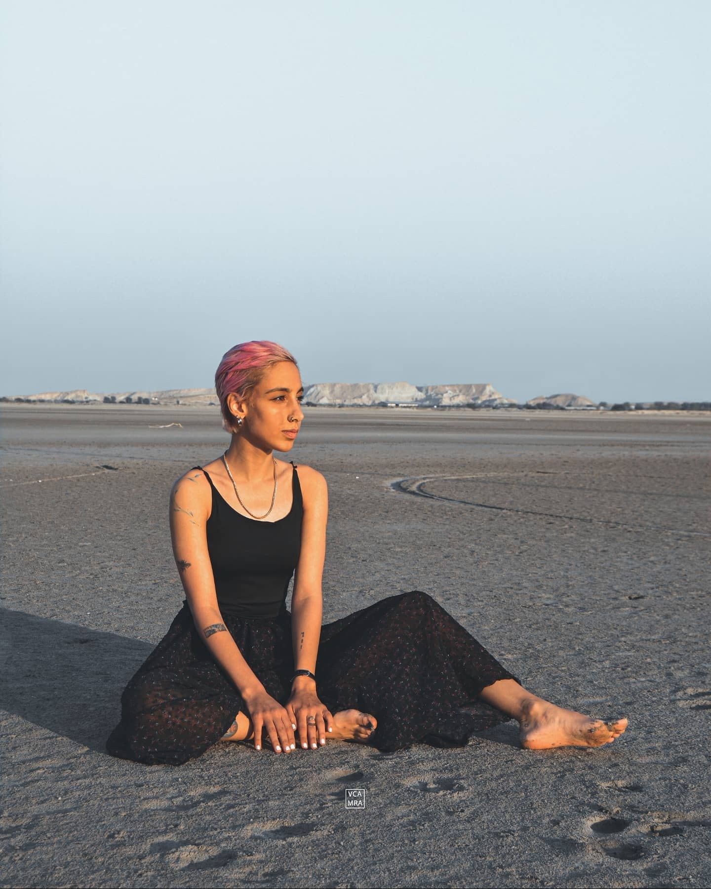 Woman with short pink hair in a black top and skirt sitting barefoot on a wide salt flat with pale cliffs on the horizon