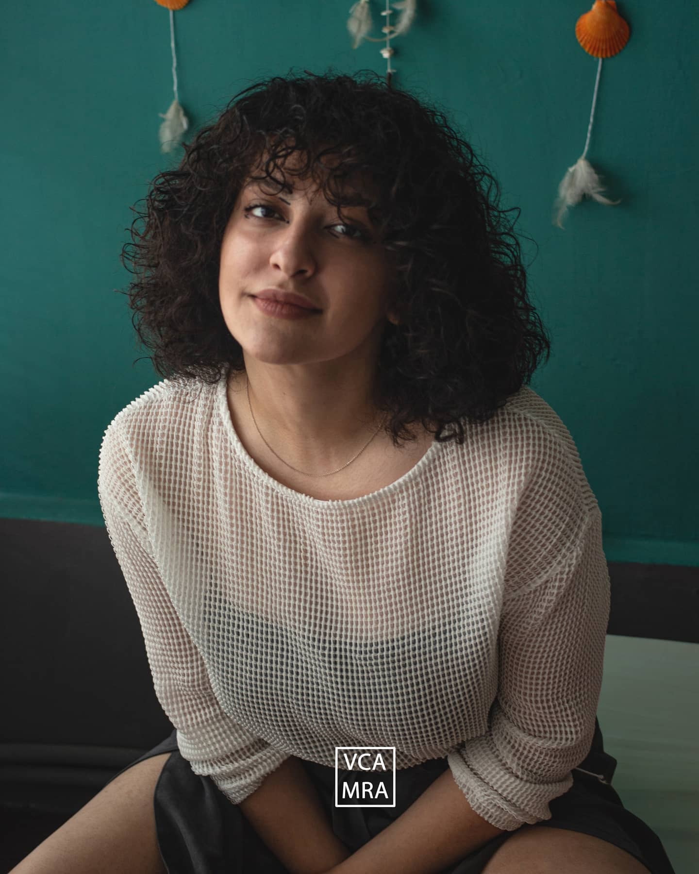 Woman with curly hair in a white open-knit sweater sitting against a teal wall hung with shells and feathers