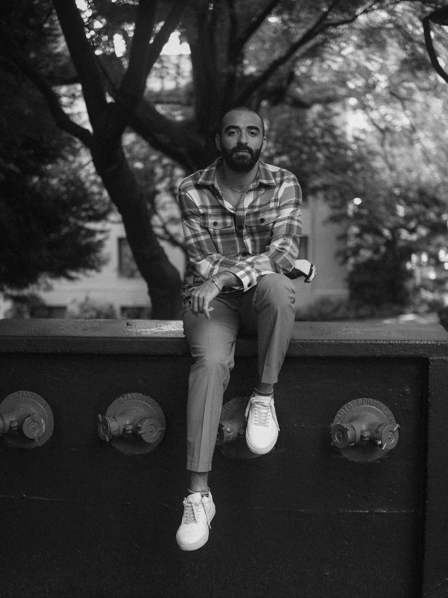 Black and white portrait of a bearded man in a plaid shirt sitting on a wall above sprinkler standpipes, trees behind him