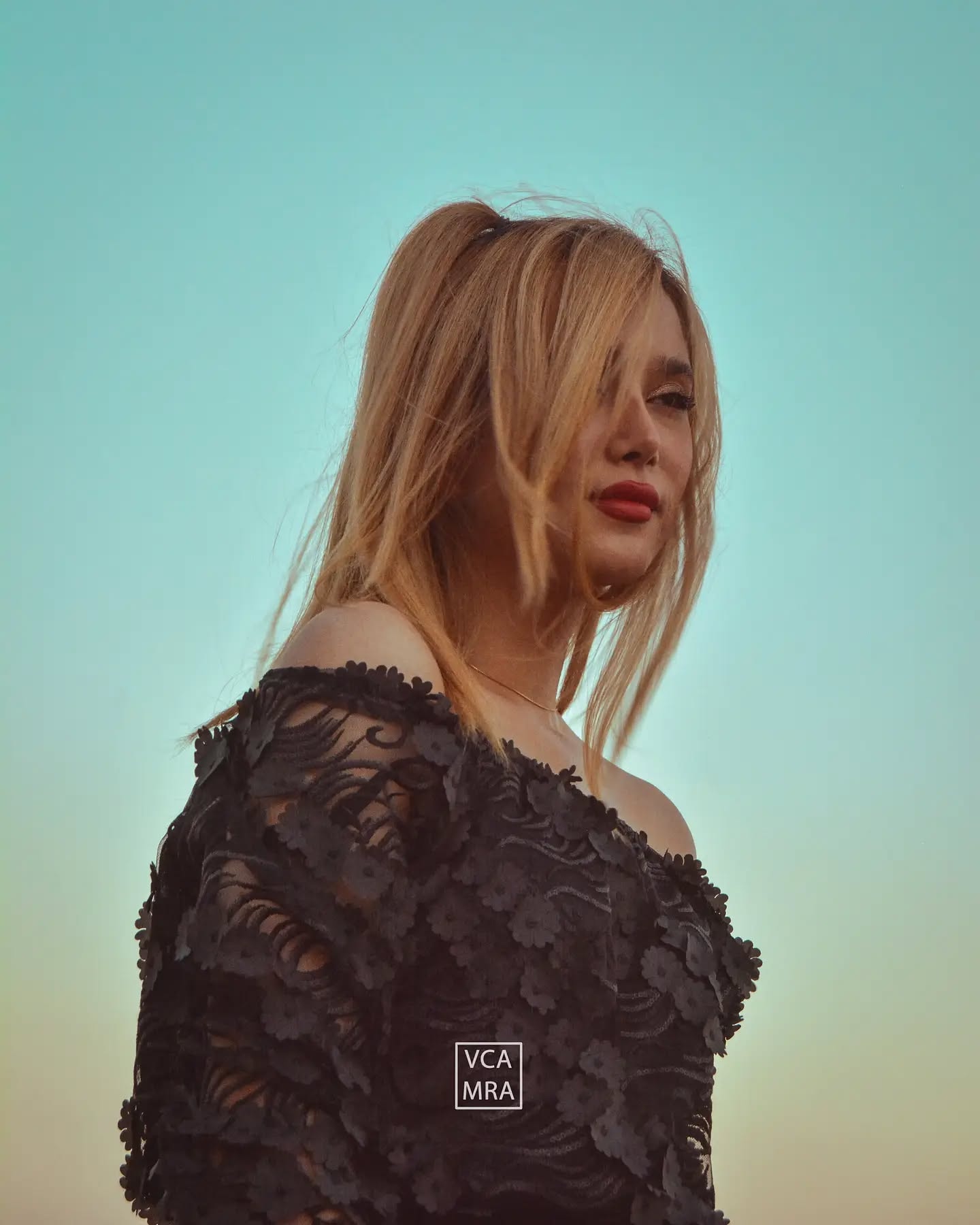 Blonde woman in an off-shoulder black floral lace top against a clear teal evening sky