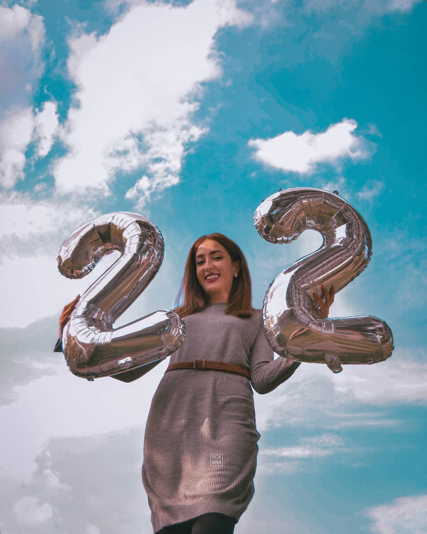 Smiling woman holding two large silver number 2 balloons against a bright blue sky with clouds