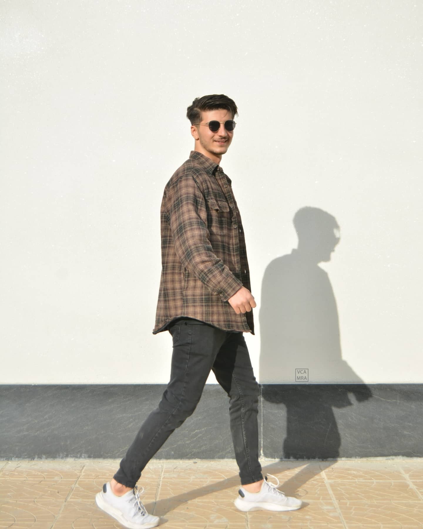 Man in sunglasses and a brown plaid shirt walking past a white wall, his shadow walking beside him