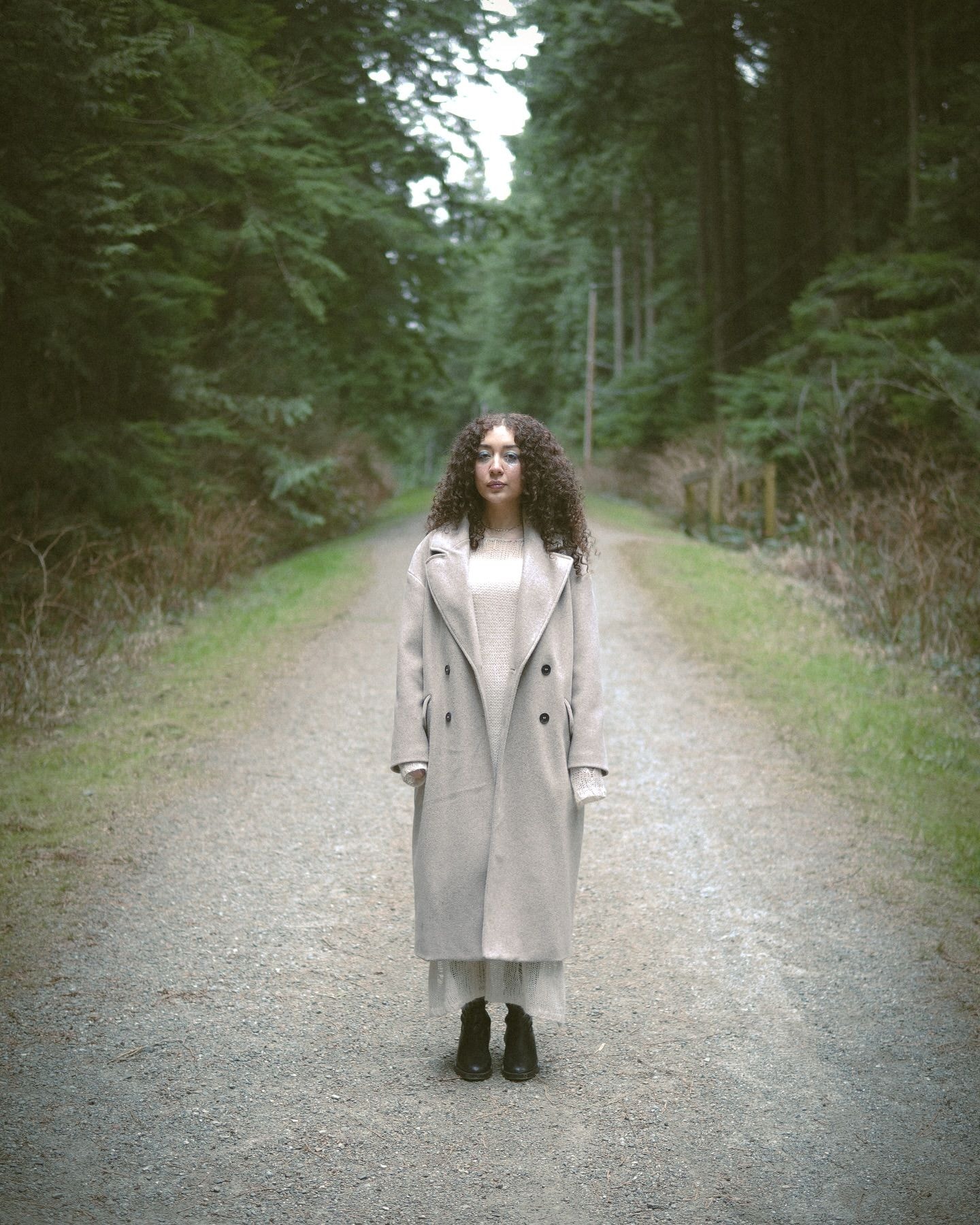 Woman with long curly hair in an oatmeal wool coat standing in the middle of a gravel forest road between tall evergreens