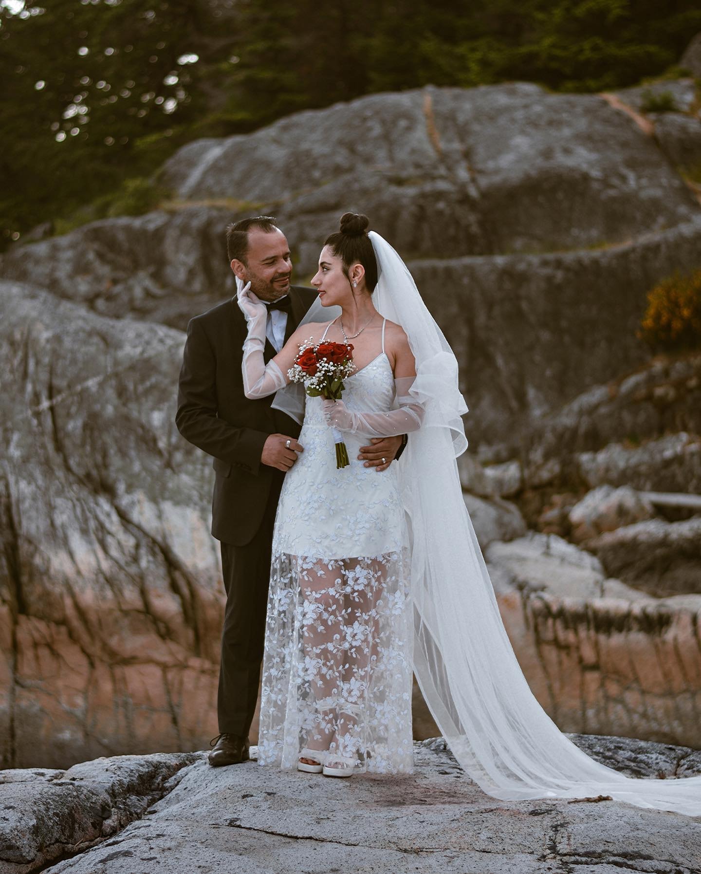 Bride in a lace gown and long veil holding red roses, touching the groom's face as they stand together on granite rocks
