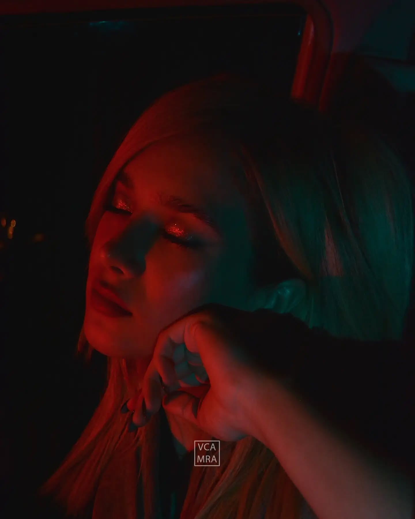 Blonde woman resting her chin on her hand with eyes closed, lit by red and teal light inside a car at night