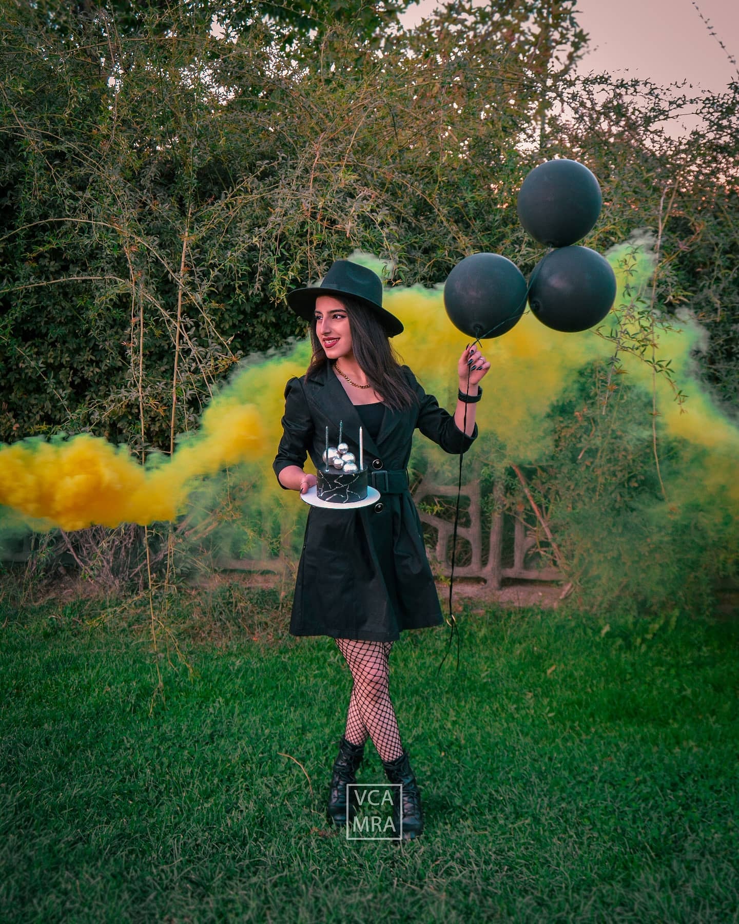 Woman in a black hat and trench dress holding a black birthday cake and black balloons as yellow and green smoke drifts behind her