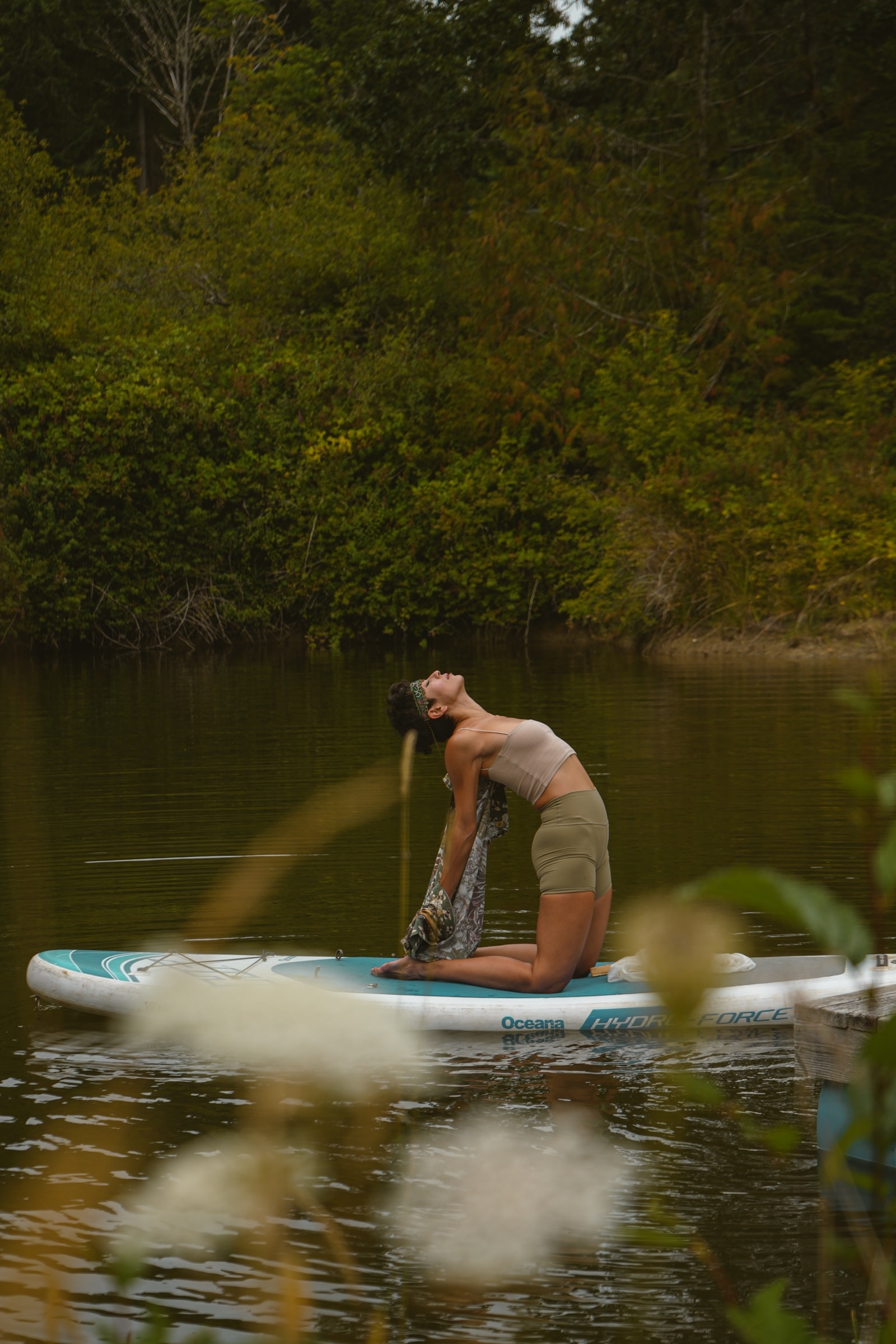 Woman arching backward on a paddleboard on a still green lake, forest behind her