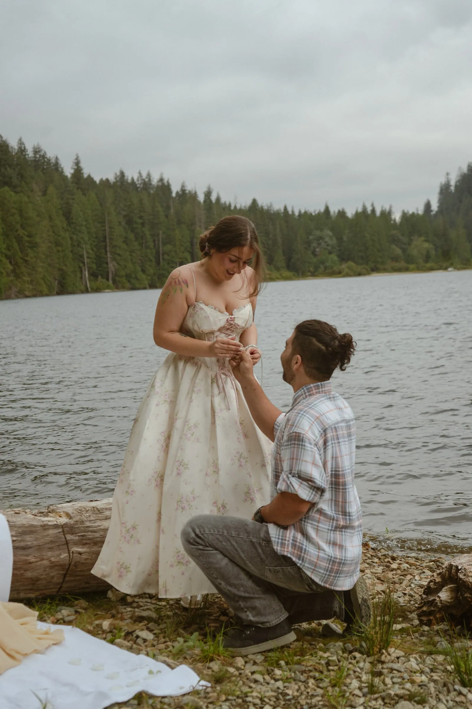 Man in a plaid shirt down on one knee on a pebbly lakeshore, sliding a ring onto the finger of a smiling woman in a floral corset dress, forested hills across the lake