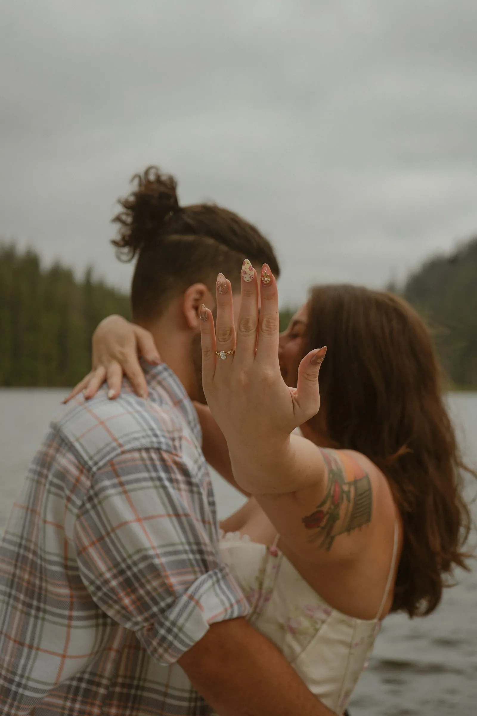 Newly engaged woman holding her hand up to the camera to show her engagement ring while kissing her partner by a lake