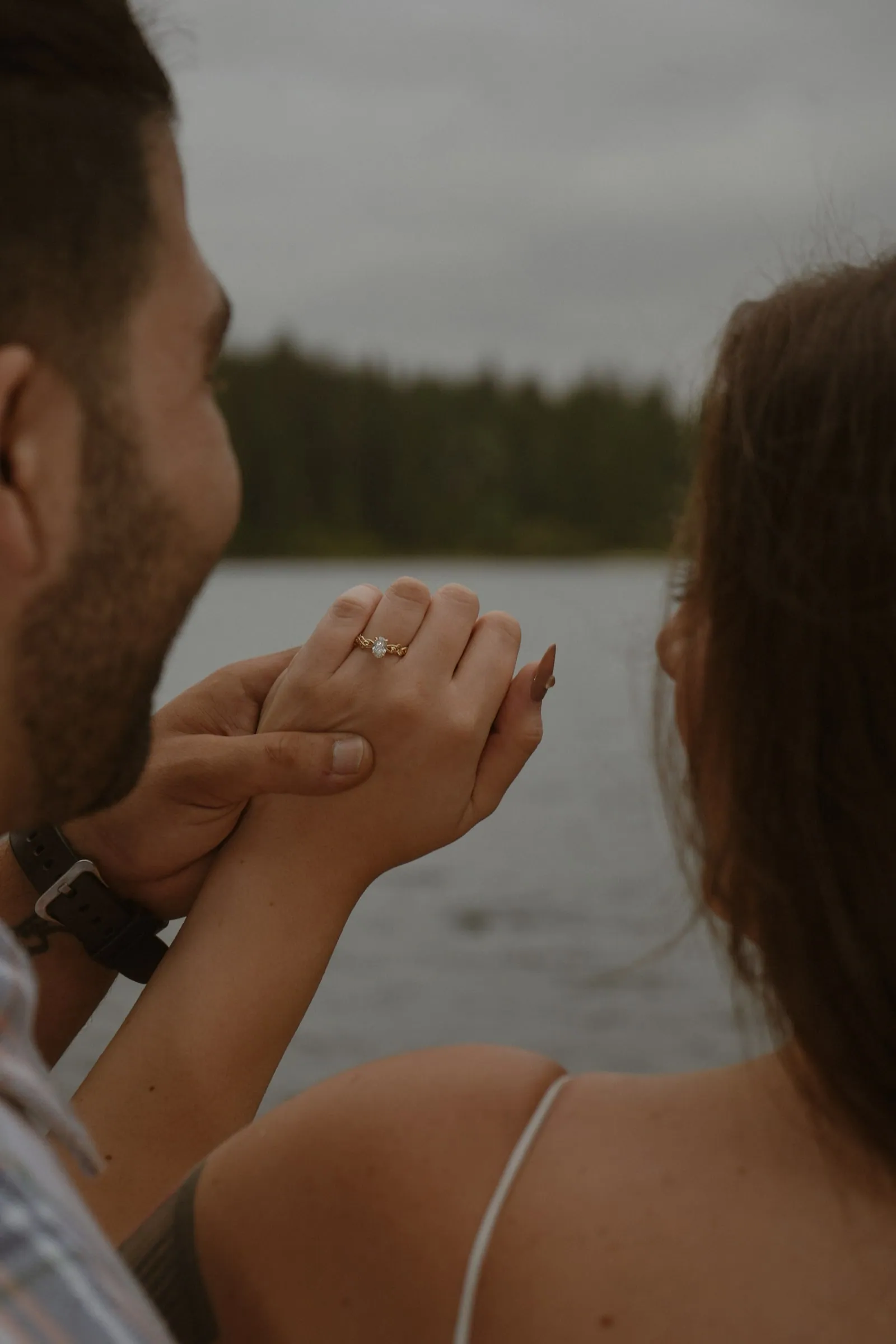 Close-up over a couple’s shoulders of a man holding a woman’s hand to show a gold engagement ring, a lake and dark forest beyond