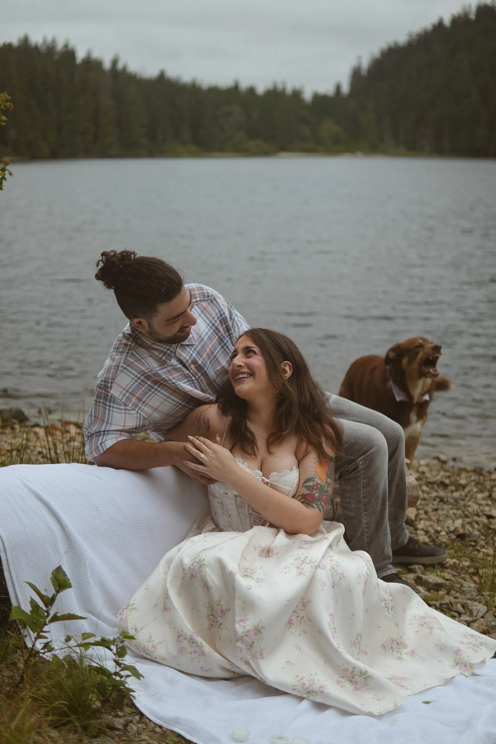 Engaged couple laughing together on a white blanket on a lakeshore while their brown dog barks beside them