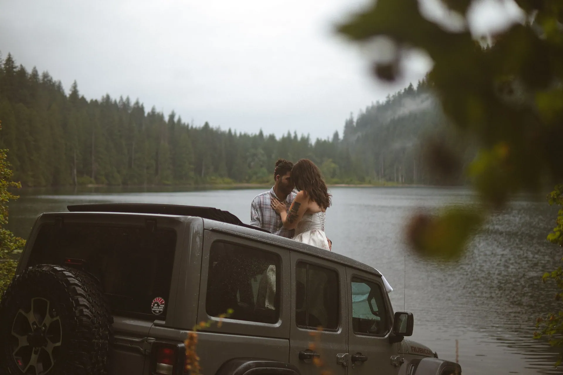 Couple embracing on the roof of a grey Jeep parked by a lake, seen through out-of-focus leaves, forest and mist on the far shore