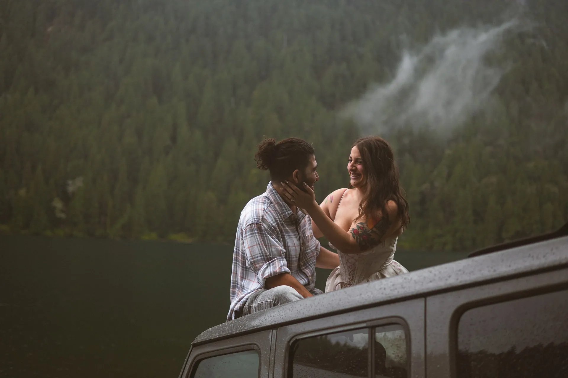 Laughing woman in a floral dress holding the face of a man in a plaid shirt as they sit on the wet roof of a Jeep, misty forested mountain behind them