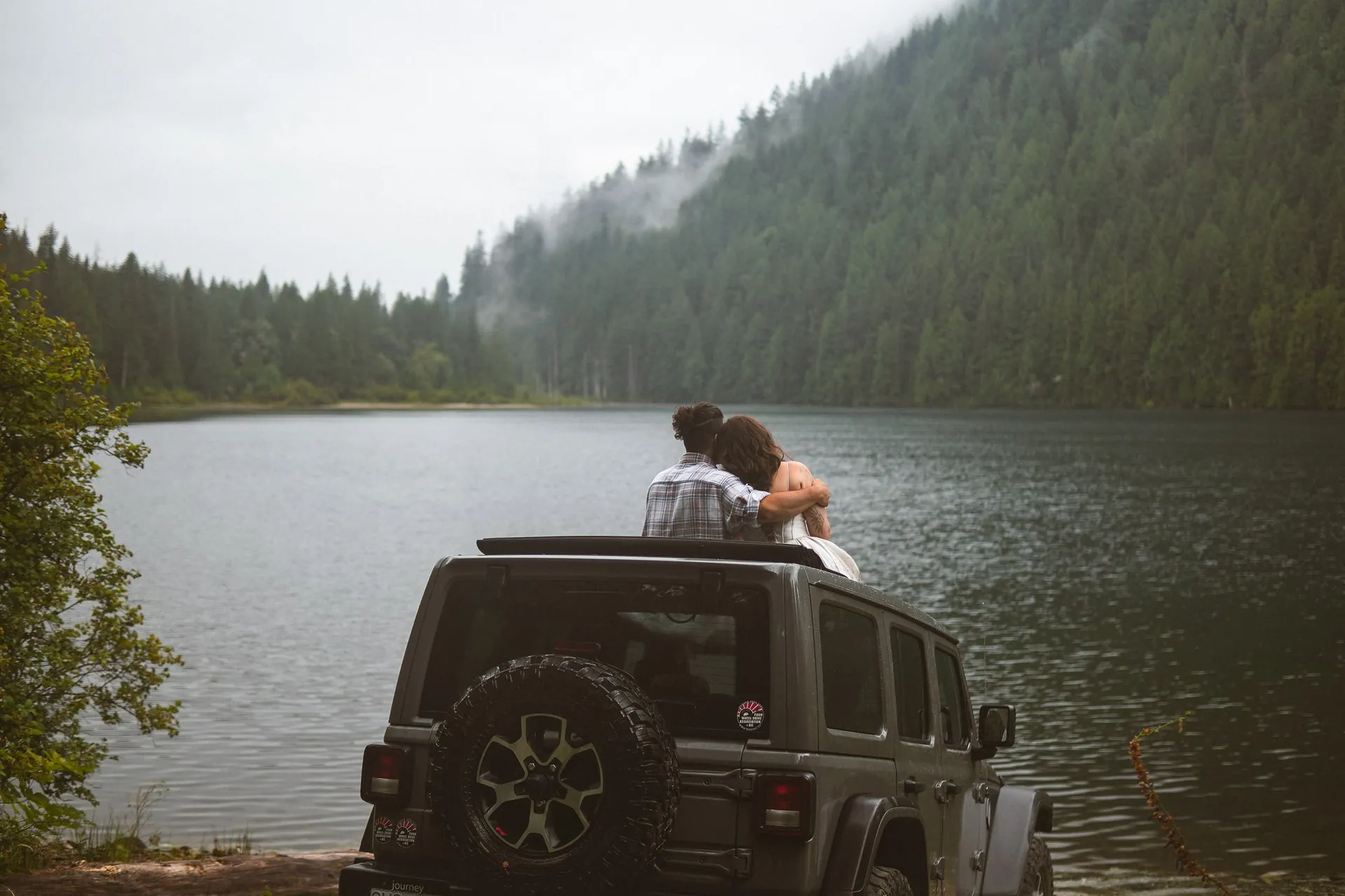 Couple seen from behind sitting arm in arm out of the open roof of a Jeep parked at the edge of a lake, mist rising off the forested mountain